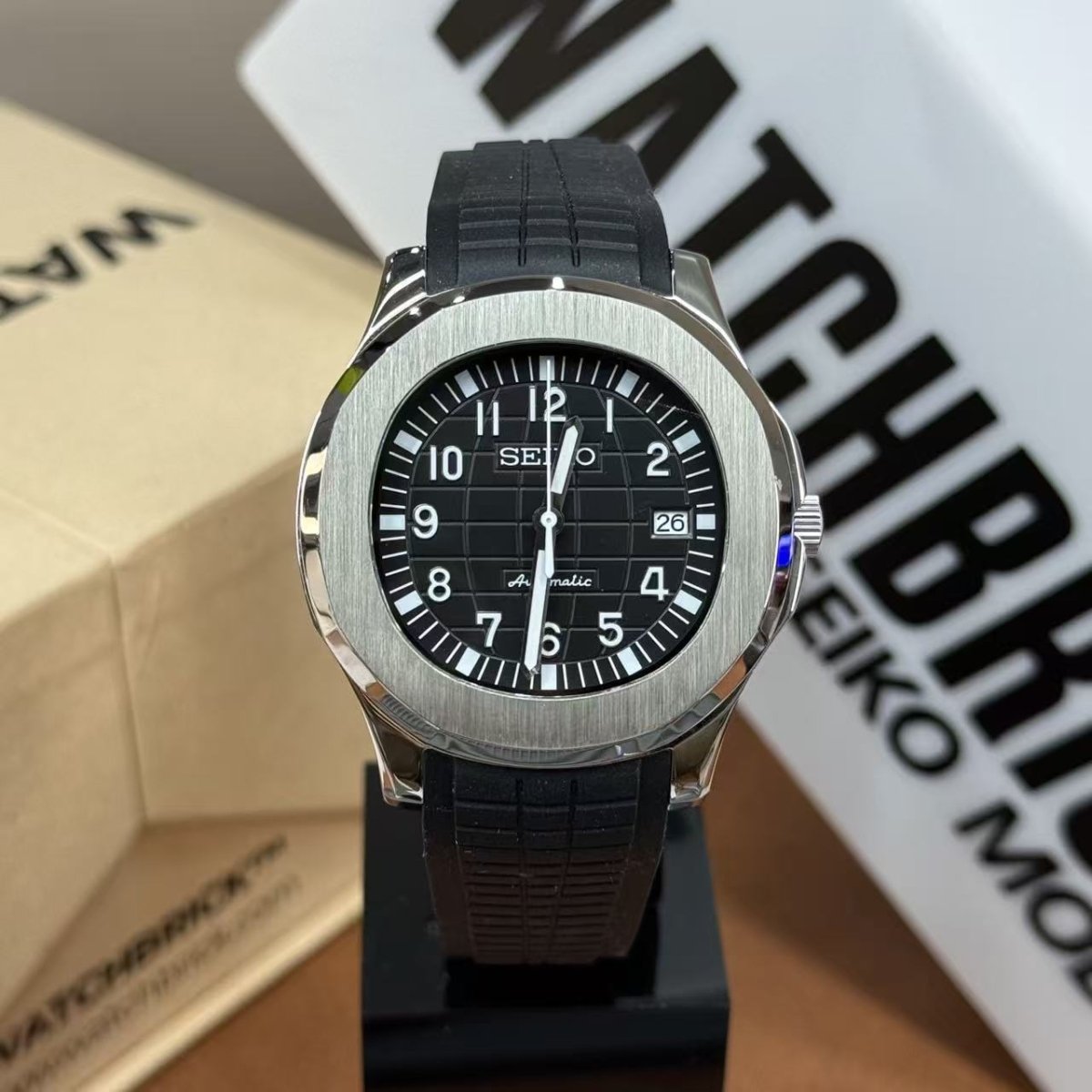 Seiko mod watch with black dial, date display, and black rubber strap on display stand