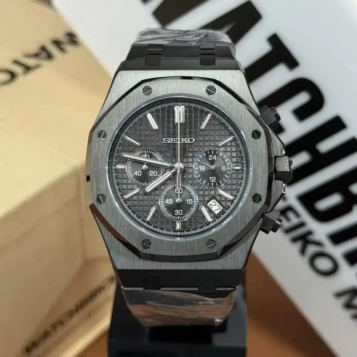 All-black Seiko-style chronograph watch with textured dial, date display, and steel bracelet.