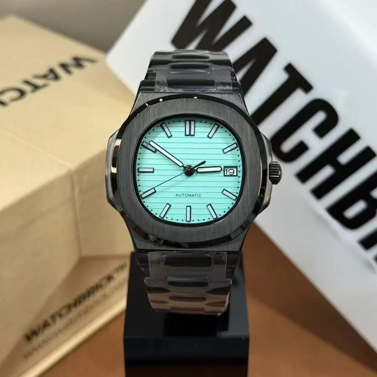 Luxury automatic wristwatch with turquoise dial, black metal case, and date window on display stand