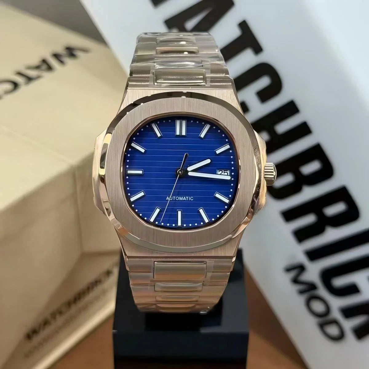 Luxury automatic watch with blue dial, silver bracelet, and date window, displayed on stand