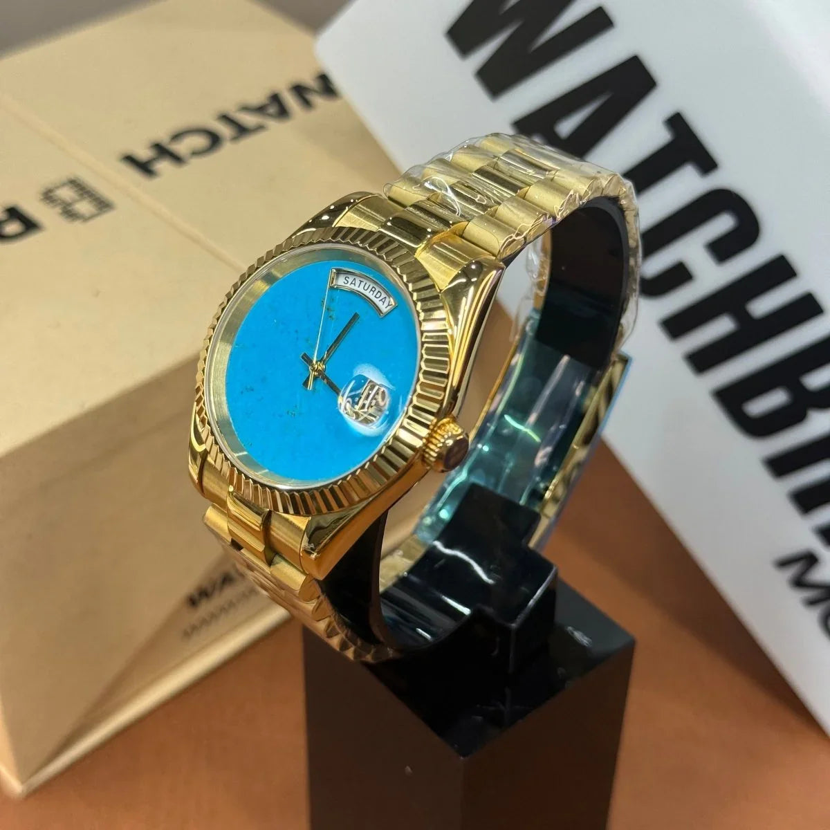 Gold mod watch with turquoise dial, day-date display, and fluted bezel on stand